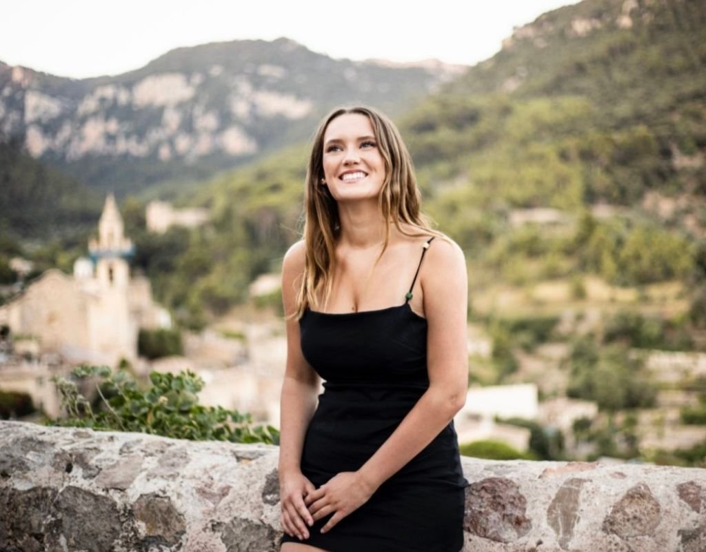 Smiling Colleen sitting on a stone wall along a road in Valdemossa, a small village in Mallorca, on a covered day. After discovering how to become a digital nomad, we started travelling and haven't looked back since.