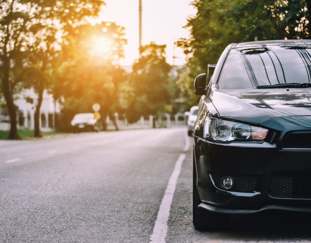 A sleek black car is parked along a tree-lined street at sunset, reflecting warm golden light. A tranquil scene reminiscent of Spanish urban charm.