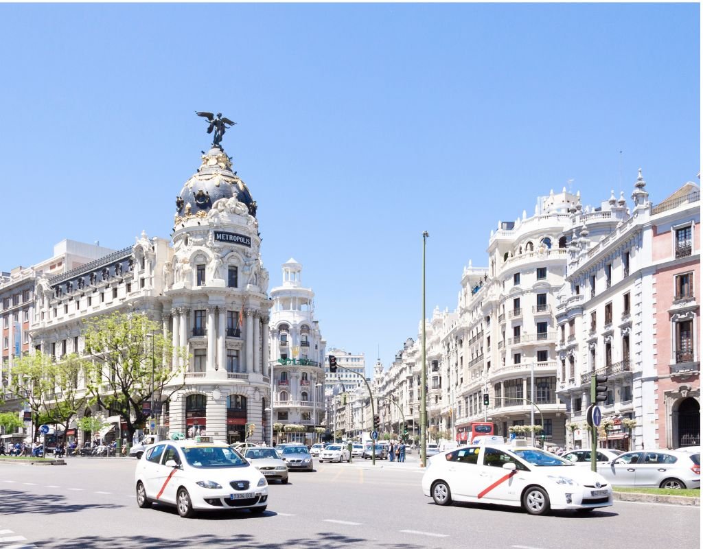 A historic European avenue with ornate buildings and white taxis passing by under a clear sky. A car in Spain can offer both convenience and style.