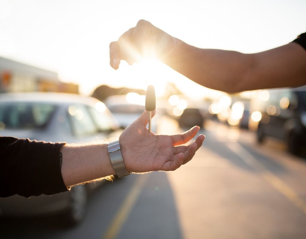 A hand holding car keys is extended to another hand in an outdoor lot, with the setting sun in the background.