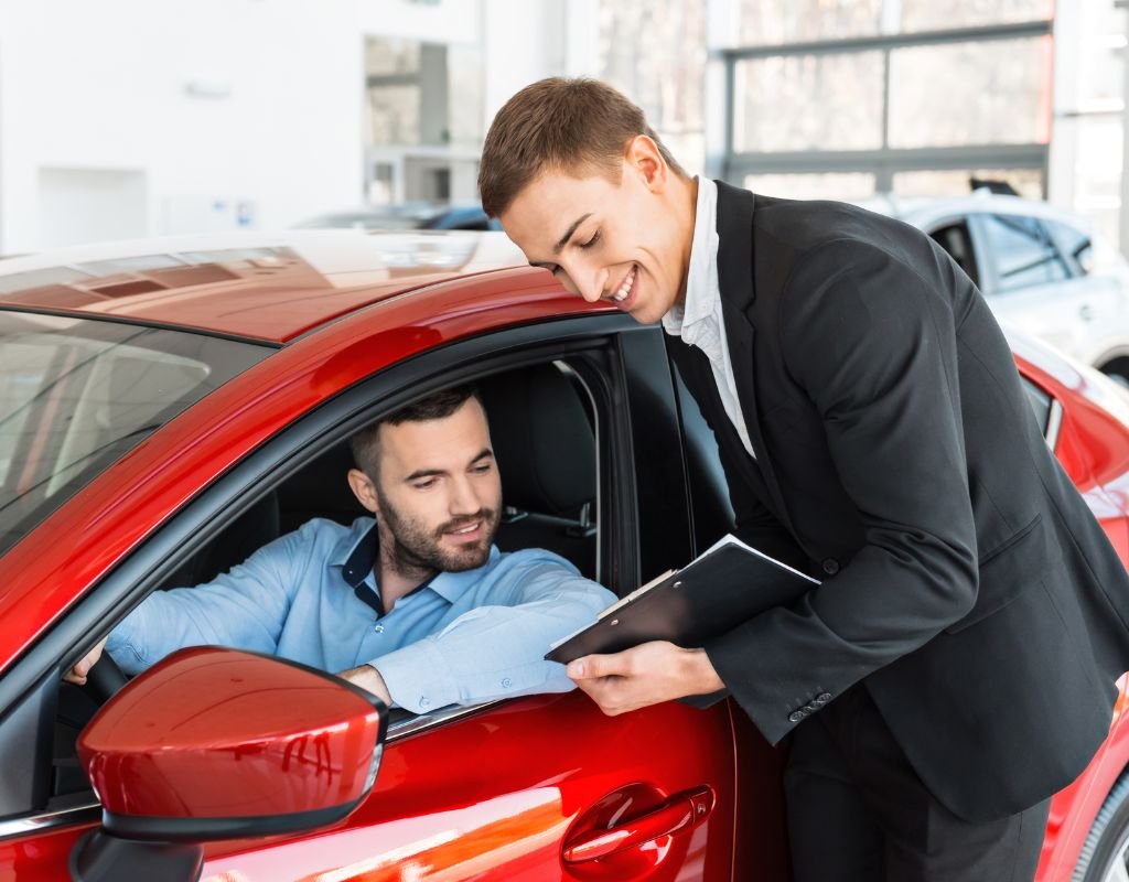 A smiling salesperson with a clipboard speaks to a man seated in a shiny red car inside a dealership. Buying a car from abroad involves consultations the customs and regulations involved.