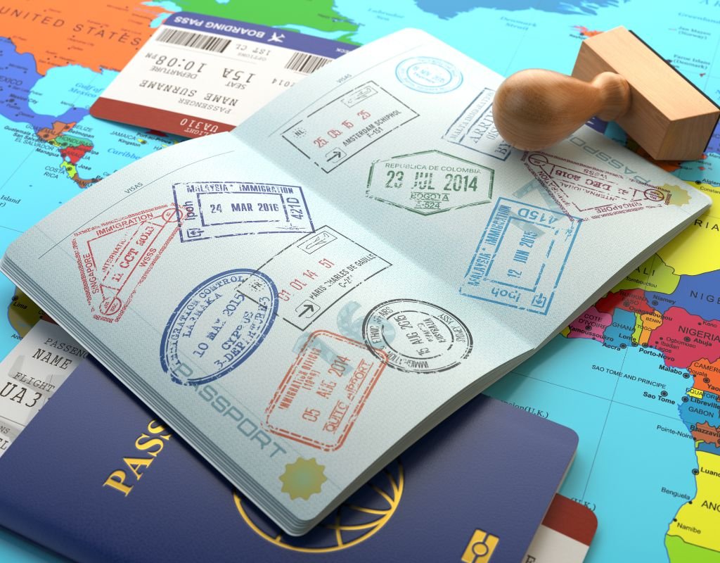 An open passport filled with colorful stamps from different countries, placed on a world map with boarding passes and a wooden stamp nearby.