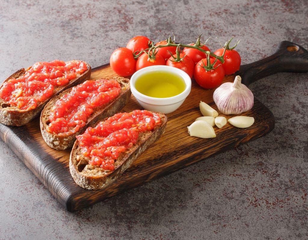 raditional Spanish pan con tomate, featuring crusty bread topped with fresh grated tomatoes, served on a wooden board alongside garlic, olive oil, and vine-ripened tomatoes. One of the simplest yet most delicious vegetarian dishes in Spain.
