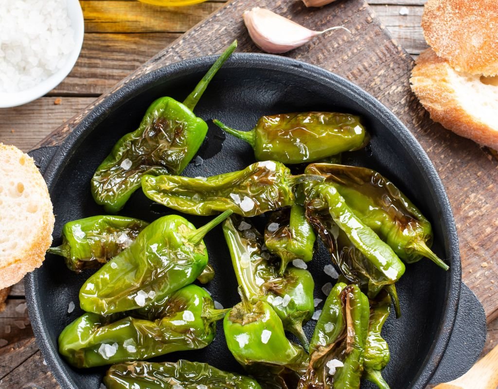 A sizzling cast-iron pan of Padrón peppers, lightly blistered and sprinkled with sea salt, served with crusty bread and olive oil—one of the classic vegetarian dishes in Spain enjoyed as a tapa.