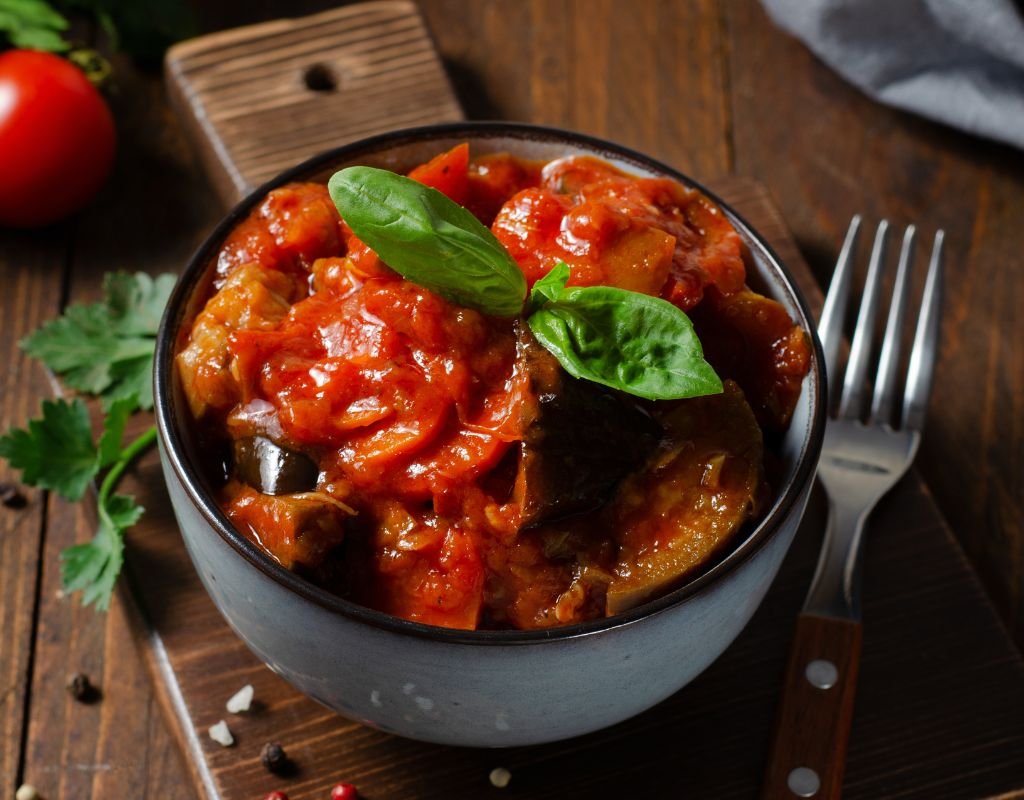 A hearty bowl of Samfaina, Spain’s version of ratatouille, made with stewed tomatoes, eggplant, zucchini, and bell peppers, garnished with a fresh basil leaf.