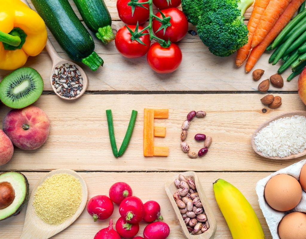 A vibrant assortment of fresh vegetables, grains, and legumes arranged on a wooden table, with the word “VEG” spelled out in green beans, carrots, and beans—celebrating the variety of plant-based ingredients used in vegetarian dishes in Spain.