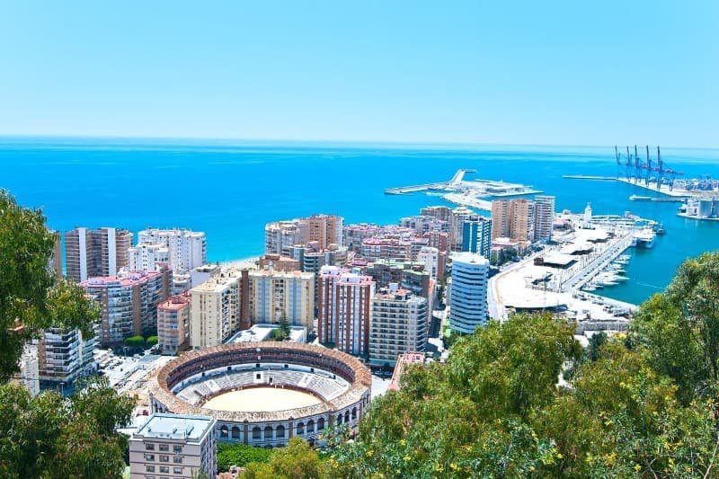 Malaga as seen from above the green trees, out to the ocean where skyscrapers are built up against the marina out on the blue ocean on a sunny day.