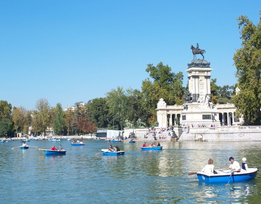 People row small blue boats on the scenic lake at El Retiro Park in Madrid, with the grand monument of Alfonso XII standing tall in the background.