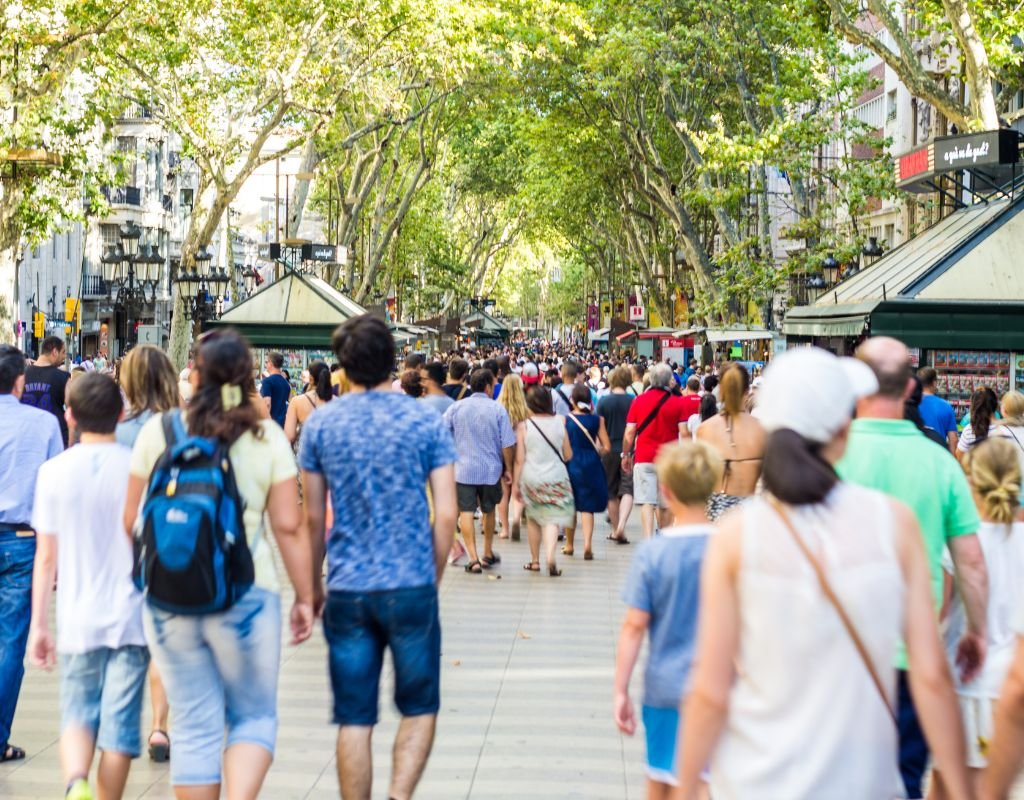 A bustling pedestrian street in Madrid, lined with lush green trees and market stalls, as crowds of tourists and locals walk along the lively boulevard.