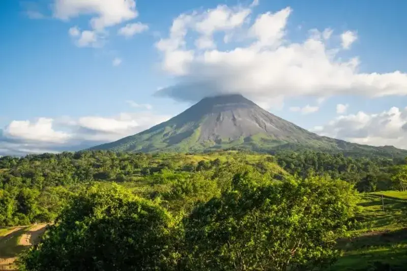 The Arenal volcano towering above the greenery on a sunny day with a few clouds at its peak.