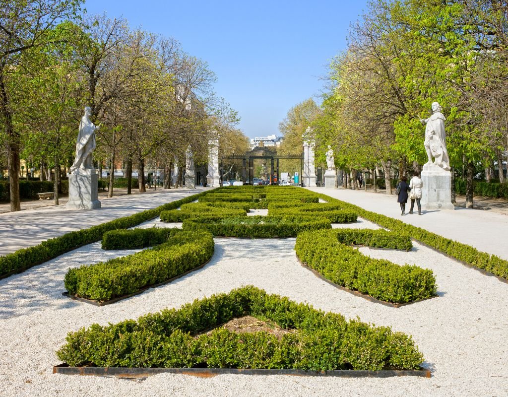 A beautifully manicured garden with trimmed hedges and marble statues in Madrid’s Retiro Park, with two people strolling under the shade of the trees.