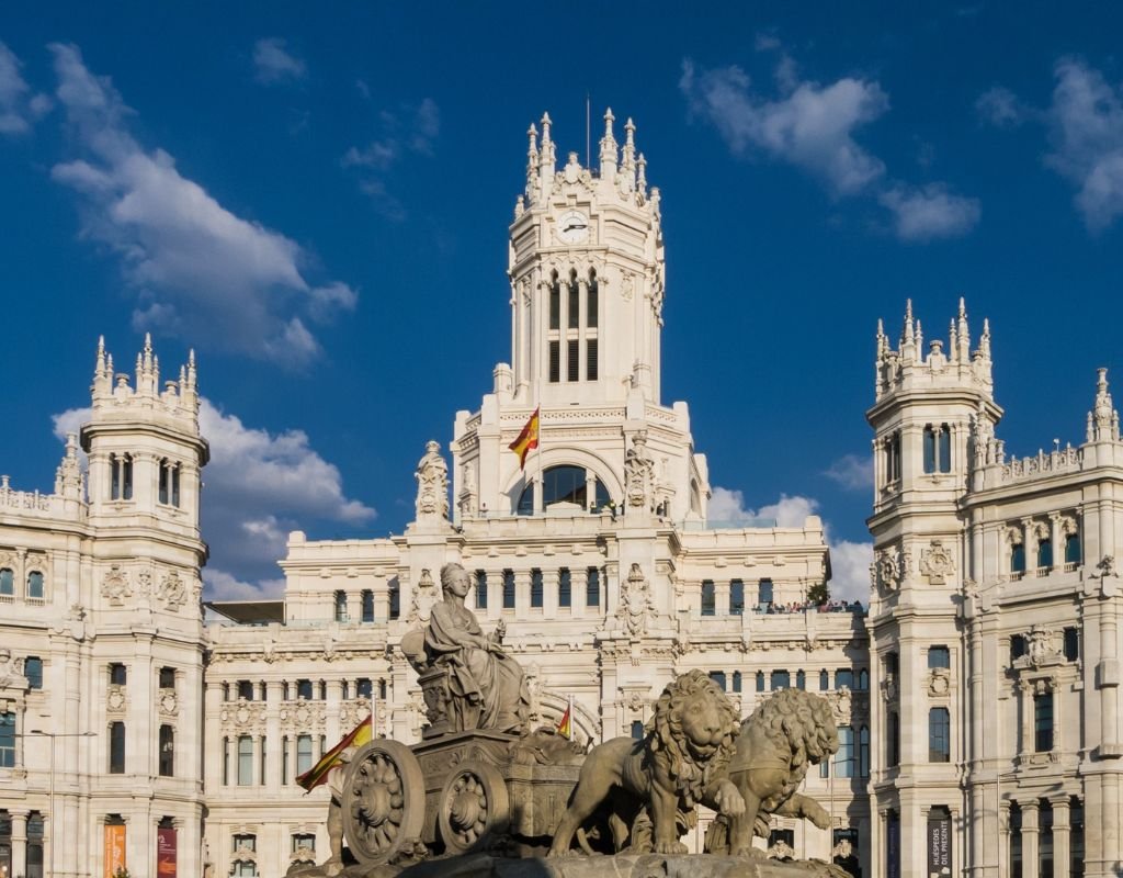 The iconic Cibeles Palace and Fountain in Madrid, with a Spanish flag flying atop the grand white building under a clear blue sky. Is Madrid worth visiting? Its impressive architecture and cultural significance make it a must-see.