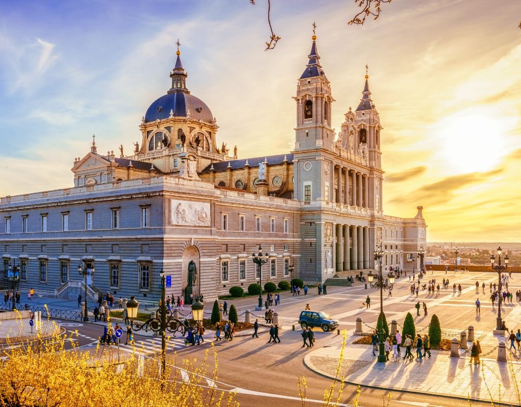 The grand Almudena Cathedral in Madrid, bathed in golden sunset light, as people walk through the spacious plaza in front of the majestic building.