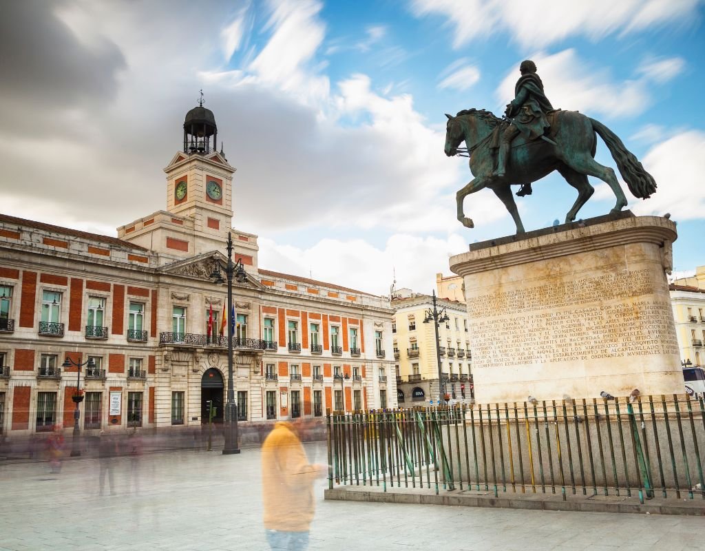 The Puerta del Sol square in Madrid, featuring the equestrian statue of King Carlos III and the historic clock tower of the Royal House of the Post Office, with people walking in the foreground.