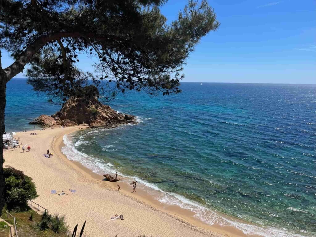 Beach in Costa Brava with white sand and an outcropping of rocks at one end of the beach.