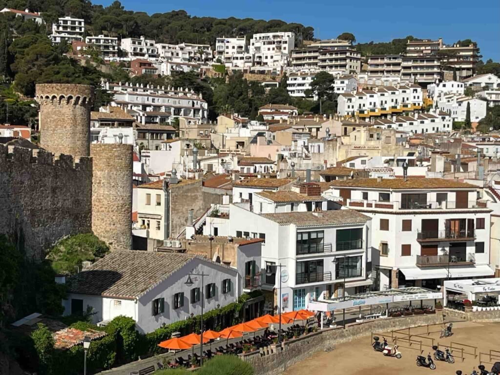 Overview of the Tossa de Mar in Spain. White buildings built up into the mountain side come all the way down to the beach, where a motor bike rental shop has many bikes parked.