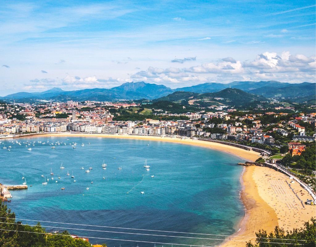A panoramic view of La Concha Bay, with golden sandy beaches, blue waters dotted with sailboats, and lush green hills in the distance. San Sebastian’s picturesque coastline offers many beautiful places to stay.