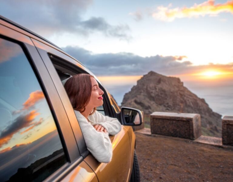 A woman leans out of a car window, smiling peacefully as she enjoys a sunset over rugged Spanish cliffs. For those considering buying a car in Spain, moments like these are easily within reach.