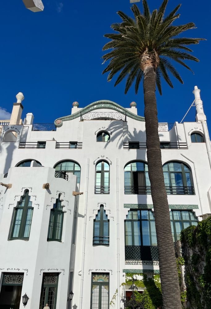 A grand white building with unique arched windows and intricate details, standing under a clear blue sky beside a tall palm tree—a striking example of Spanish architectural style.