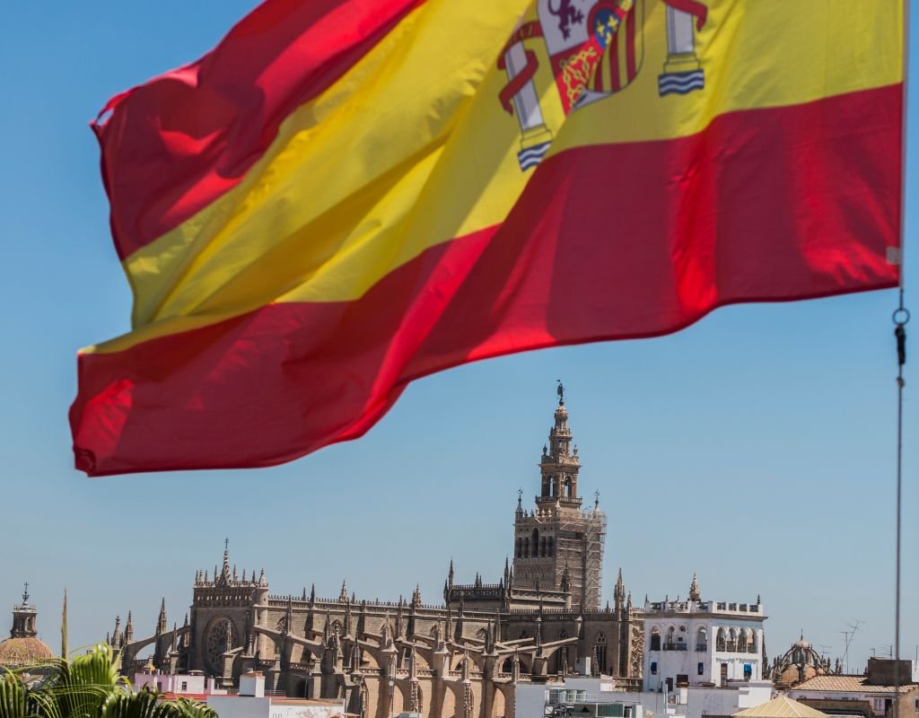 The Spanish flag waves proudly in the foreground with Seville's famous Giralda tower in the background. To start the process of getting Spanish citizenship, a connection to Spain’s culture and language is essential.