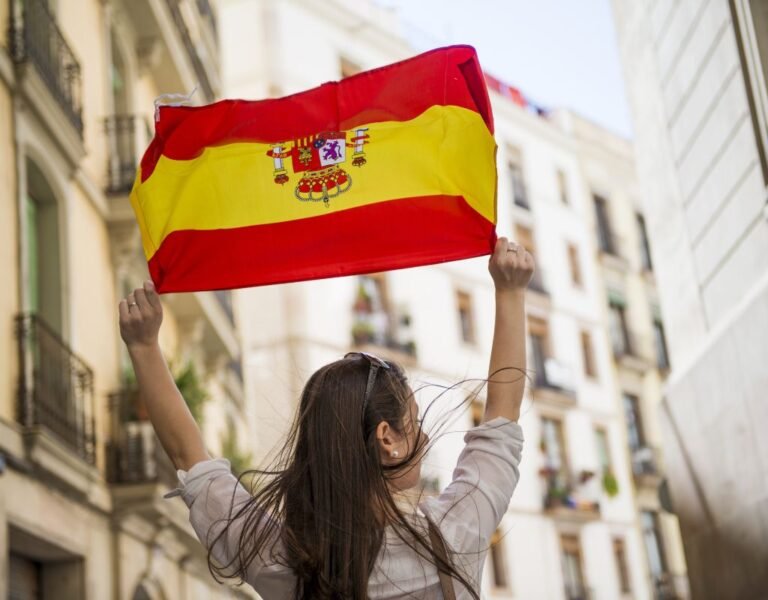 A person raises the Spanish flag in a lively street scene, celebrating Spain’s national pride. How to get Spanish citizenship? Develop ties to the country through residency and cultural integration.