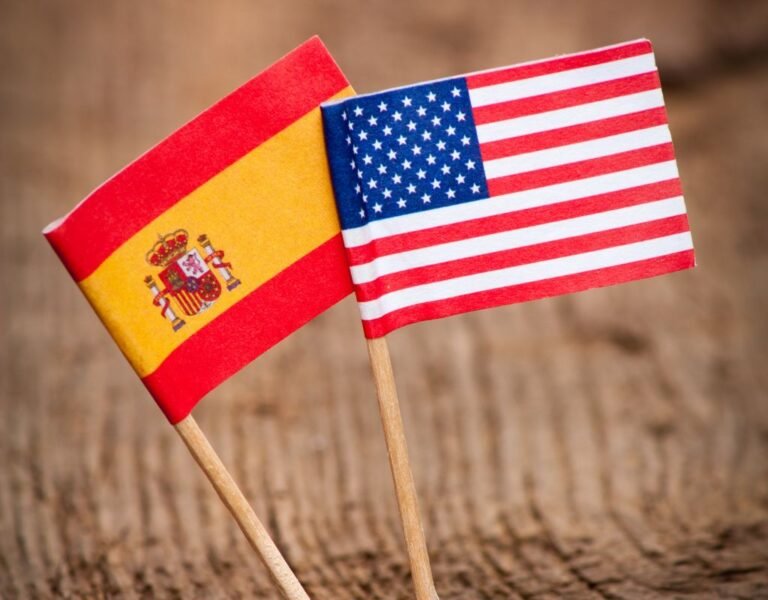 Miniature flags of Spain and the USA stand side by side on a rustic wooden table, representing cultural and financial contrasts, including cost of living in Spain vs USA.