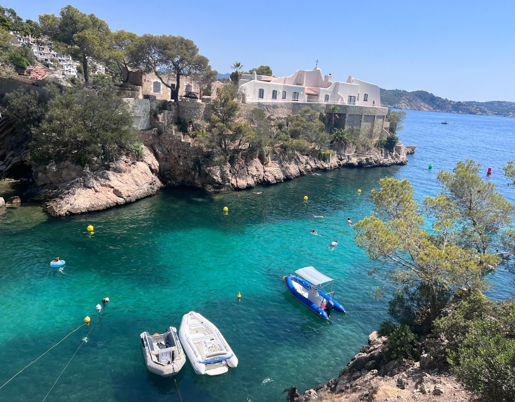 A quiet bay with crystal-clear turquoise water, small boats anchored near rocky cliffs, and swimmers enjoying the serene atmosphere, highlighting Spain’s coastal allure.