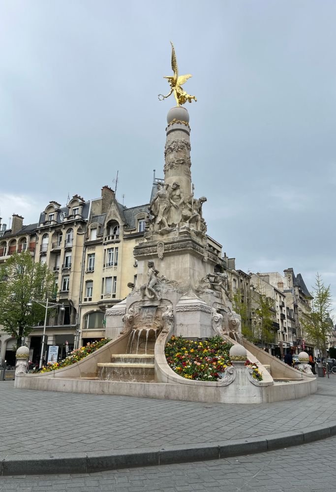 An ornate fountain in a French square with detailed sculptures and vibrant flowers at its base. Despite their beauty, squares like this are places prone to pickpockets, touching on bad things about living in France.