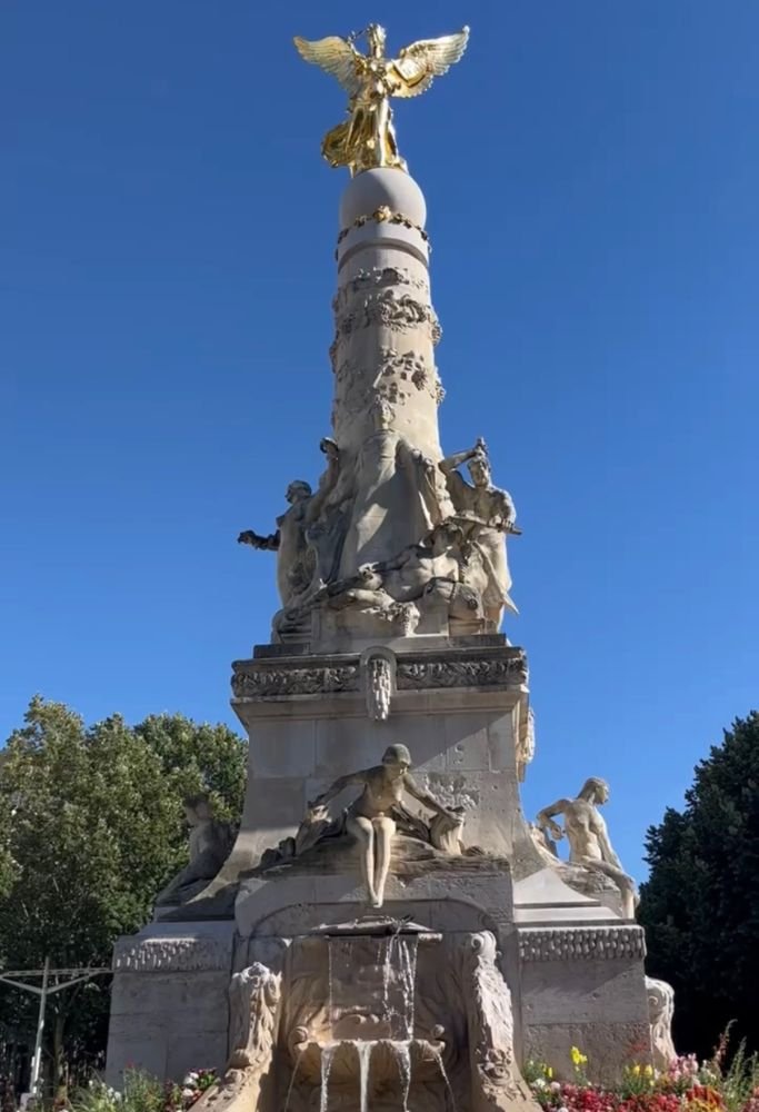 A striking statue with a golden angel atop a tall monument surrounded by intricate stone carvings and fountains, set against a vibrant blue sky. The grandeur of public art highlights the historical charm often experienced while living in France as an American.