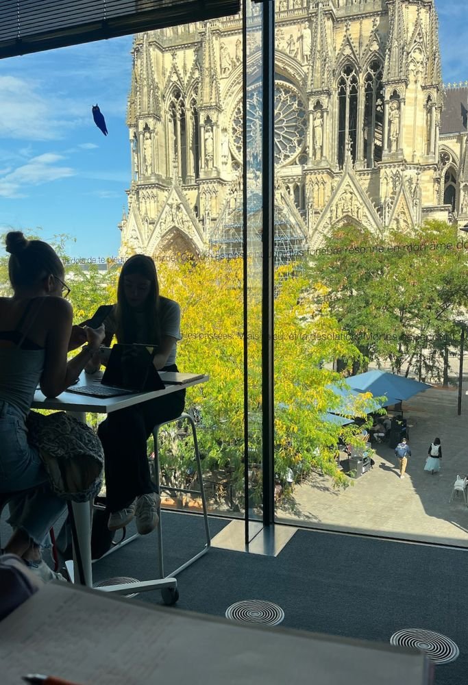 A modern study space with large glass windows framing the view of Reims Cathedral, students working inside, and the autumn landscape outside.