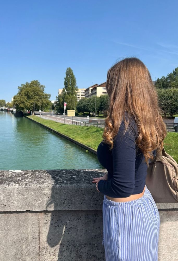 A young woman gazes over a quiet canal lined with greenery and modern buildings, enjoying a sunny day. For those living in France as an American, such tranquil urban scenes offer a unique mix of relaxation and exploration.