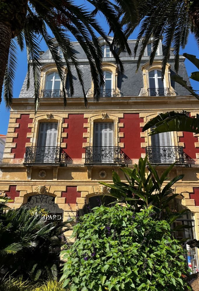 A beautifully preserved building with classic red and gold architecture, surrounded by lush tropical plants under a clear blue sky. Such views showcase the mix of historical charm and natural beauty that define living in France.