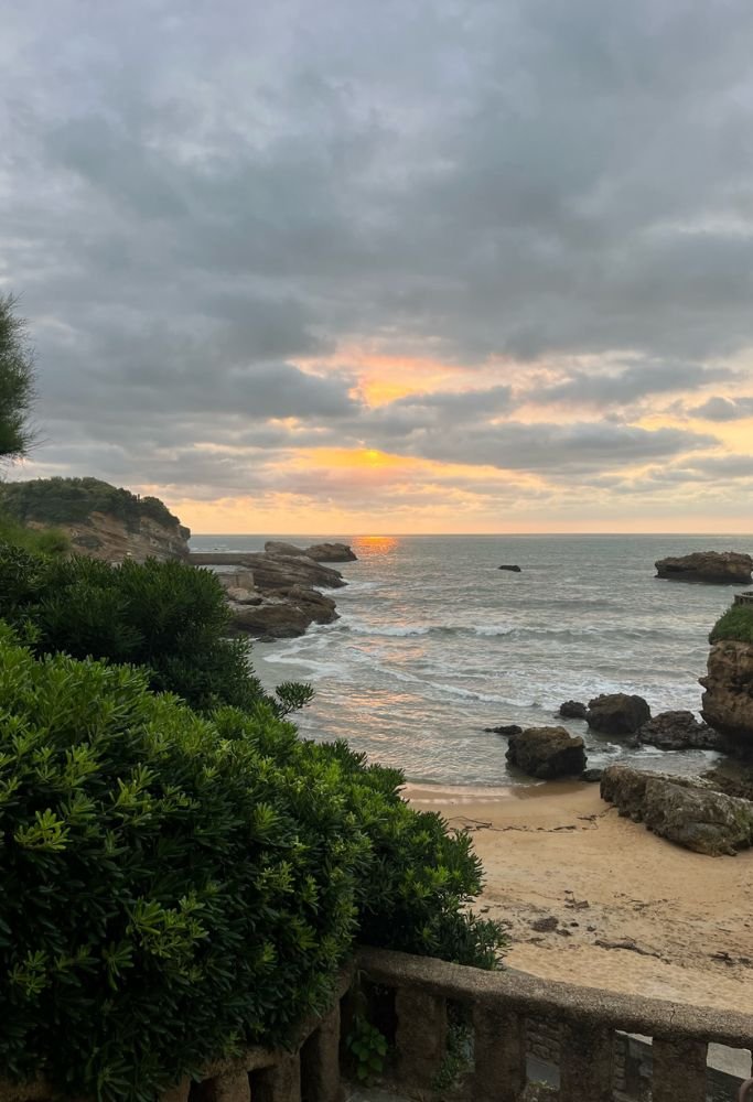 A serene coastal view at sunset with waves gently hitting the sandy shore, surrounded by lush green plants and rocky formations, evoking a peaceful moment by the ocean. Living in France as an American often brings moments of awe like this.