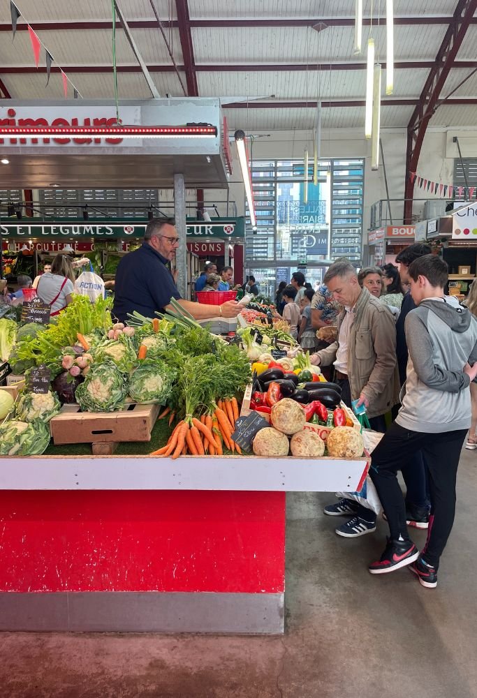 A vibrant French indoor market with fresh produce like carrots, cabbage, and peppers on a stall, with locals and tourists shopping for ingredients.