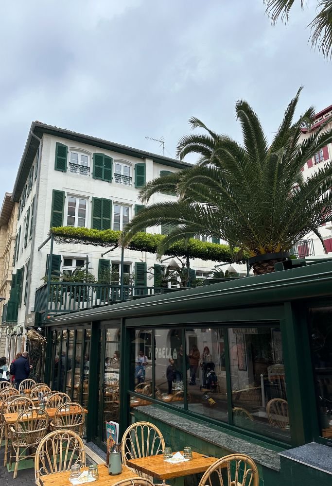 A quaint French restaurant with outdoor patio seating, featuring green window shutters, palm trees, and tables set for a sunny meal. For those living in France as an American, enjoying meals in charming settings like this is a delightful experience.