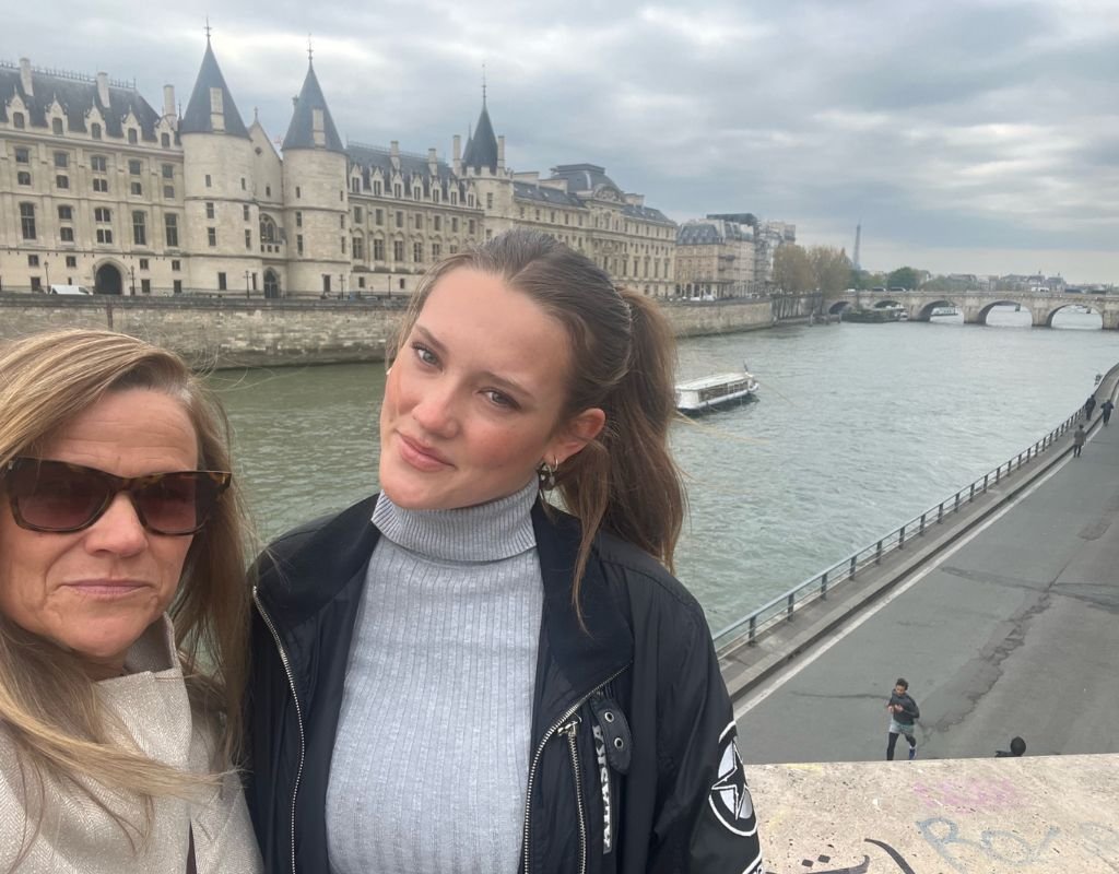 Colleen and her eldest daughter smiling on a Parisian bridge with the Seine River and historic architecture in the background on a cloudy day.