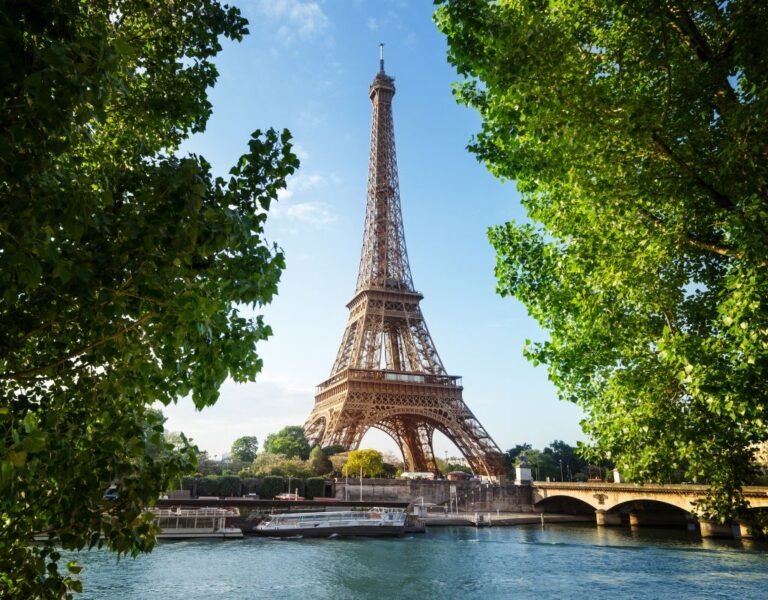 A picture-perfect view of the Eiffel Tower framed by lush green trees alongside the Seine River, illustrating a timeless Parisian scene. Living in France as an American, seeing the Eiffel Tower never loses its charm.