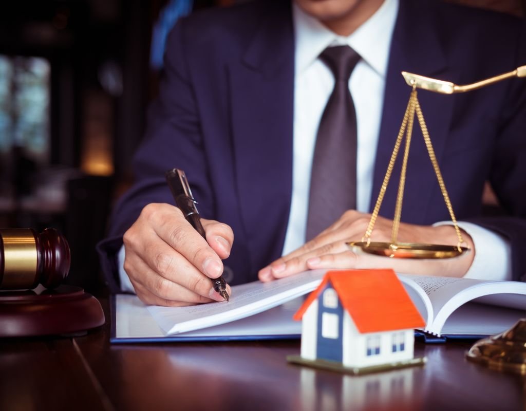 A lawyer in a suit signing legal documents at a desk, with a miniature house model and a justice scale, emphasizing the importance of legal advice when buying a home in Mexico.