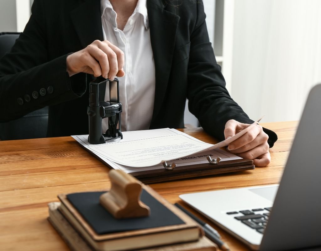 A professional in formal attire stamps a contract at a desk, surrounded by office supplies and a laptop, demonstrating the verification and approval process