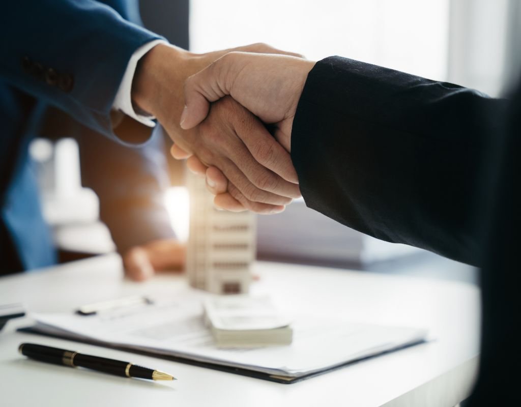 Two business professionals shaking hands in an office setting, with paperwork, a pen, and a house model in the background, highlighting a final agreement stage