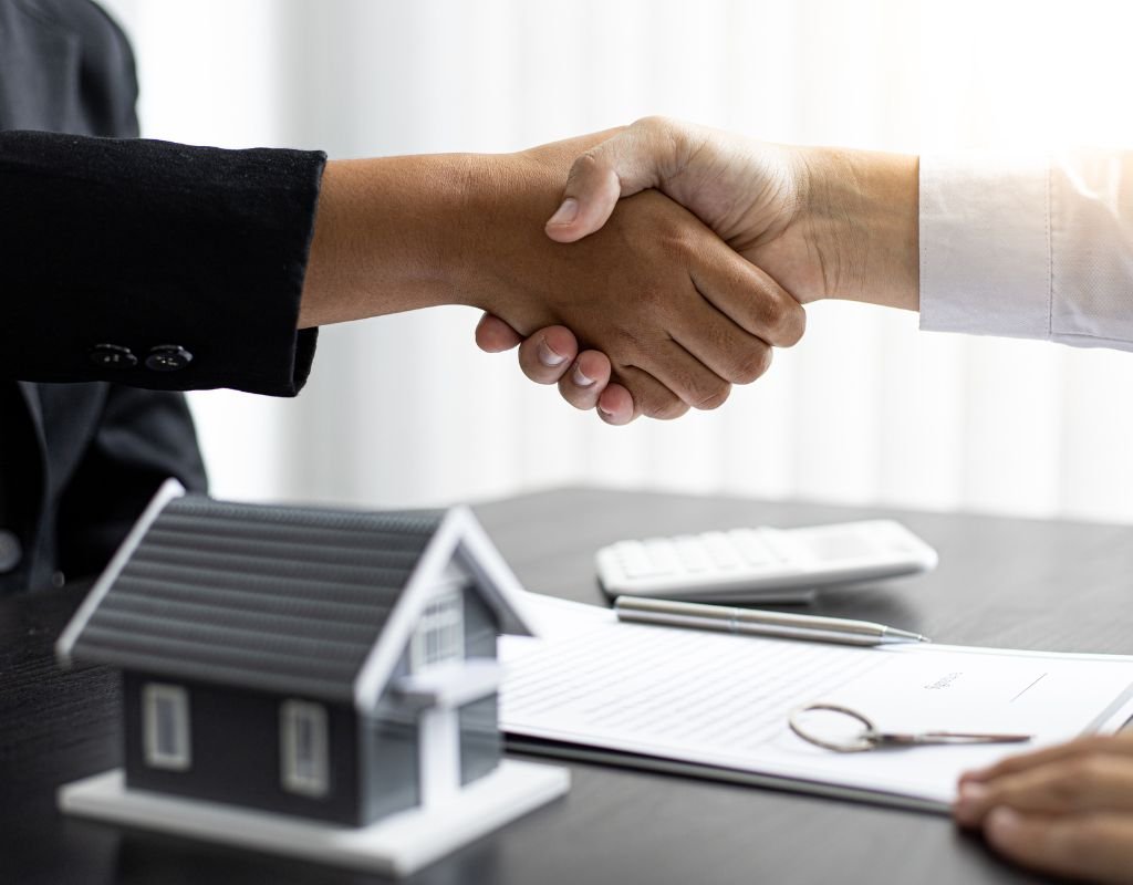 Two people shaking hands over a contract with a miniature house model on the table, symbolizing a successful real estate deal. A crucial step when buying a home in Mexico involves agreements and paperwork to finalize the purchase.