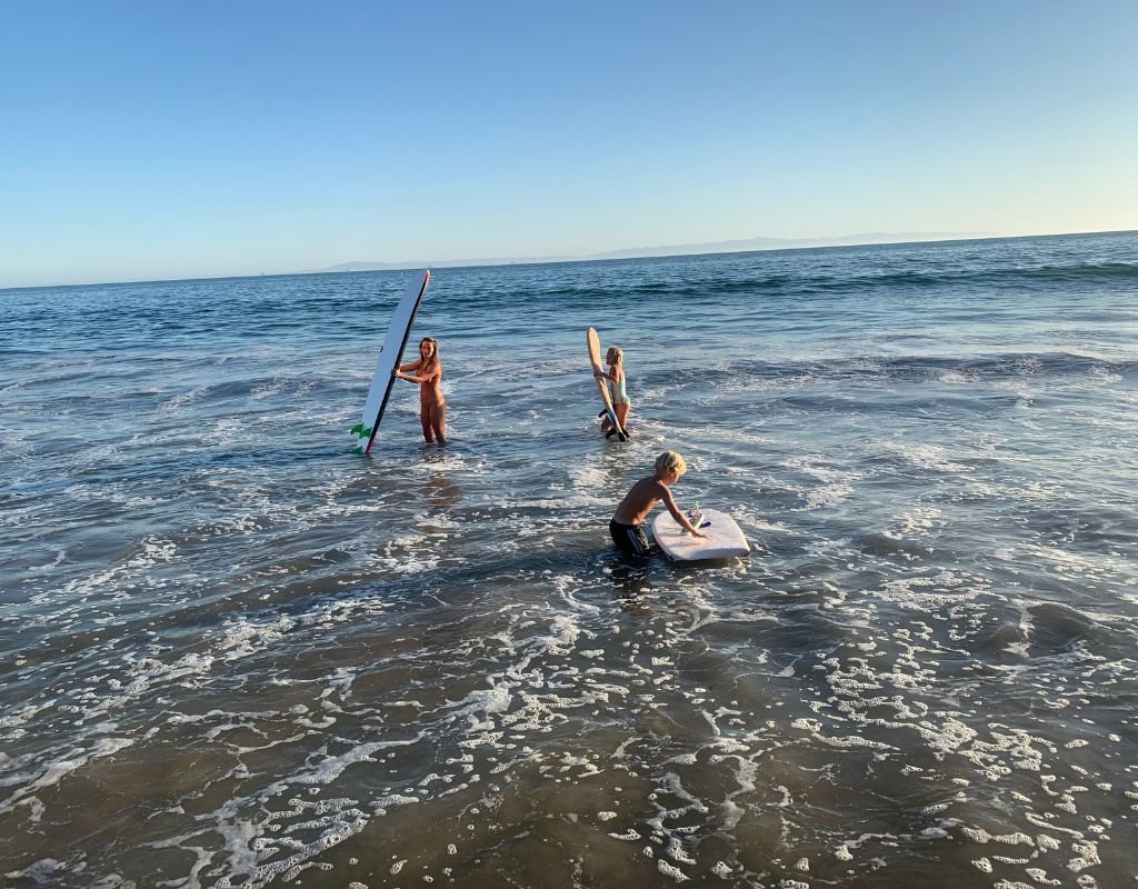 Three children playing in the ocean with surfboards under a clear blue sky, enjoying the calm waves on a sunny day.