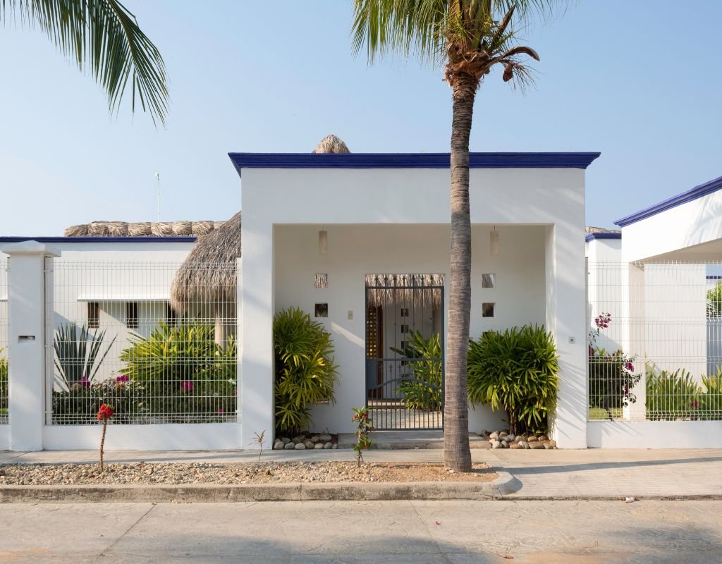 A modern white house with blue trim, tropical plants, and a palm tree in front, showcasing coastal living options available in Mexico.