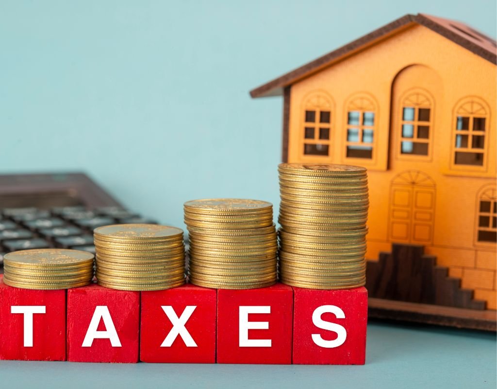 Stacks of coins on red blocks spelling "TAXES," with a wooden house model in the background, emphasizing the financial considerations and tax implications of buying a home in Mexico.