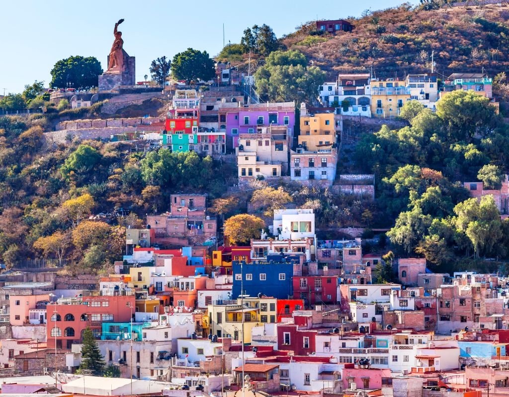 A hillside town with colorful houses stacked along the slopes, with a large statue at the top, reflecting the charm and culture that attract people to Mexico.