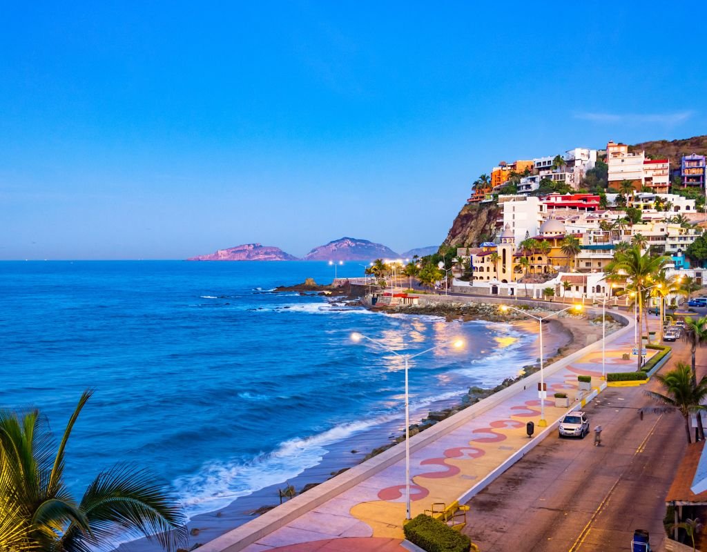 A coastal view at sunset with colorful buildings perched on a hillside, palm trees, and a scenic oceanfront walkway in Mazatlán, one of the many countries Americans can move to for a vibrant lifestyle abroad.