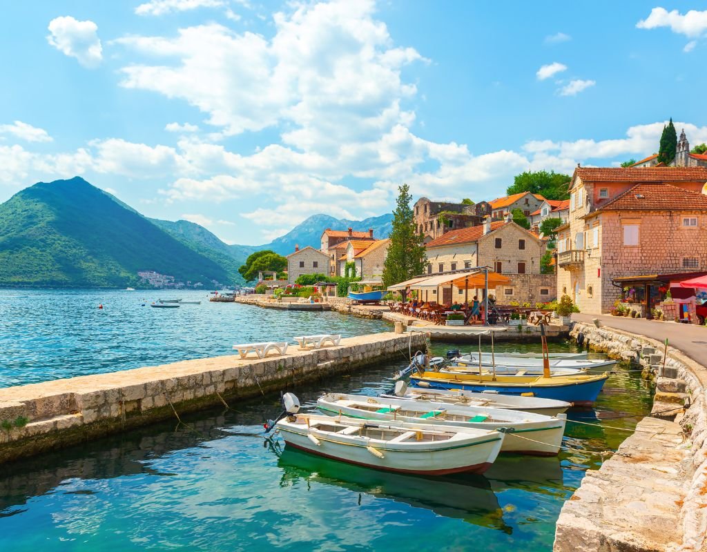 Charming stone houses with terracotta roofs line a peaceful harbor, where small fishing boats float on calm blue water in the scenic European coastal village of Montenegro.