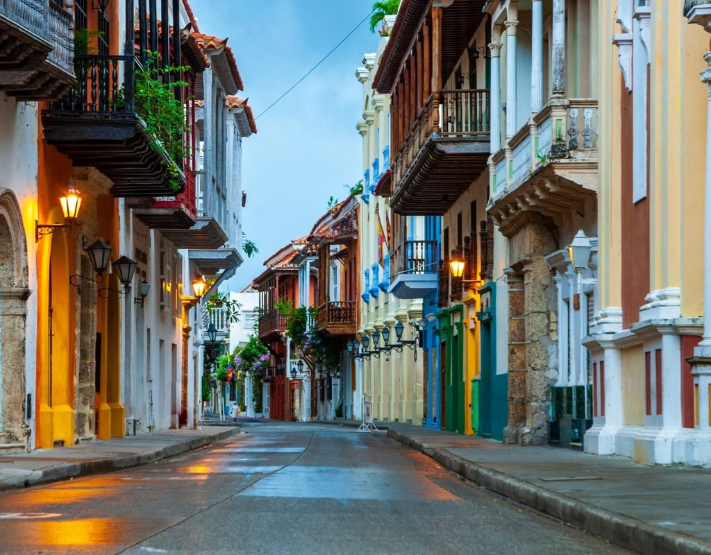 A quiet, colorful street in Cartagena, Colombia, lined with colonial-style buildings, wooden balconies, and vintage street lamps glowing in the early evening light.