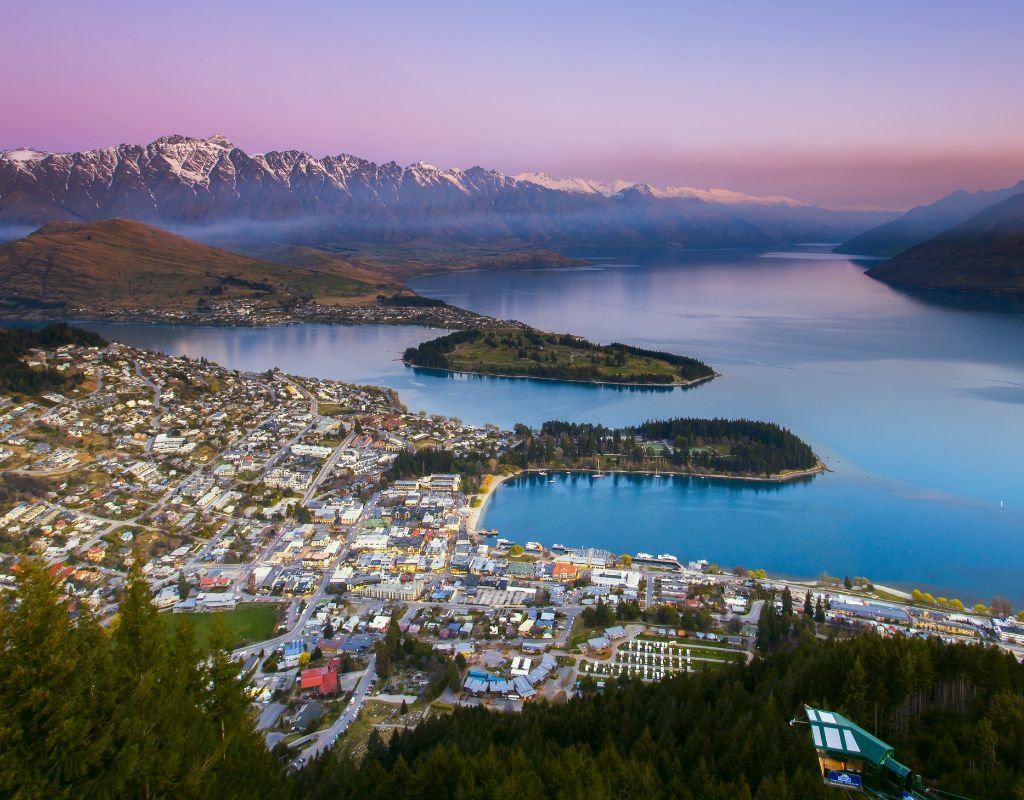 A panoramic view of Queenstown, New Zealand, with snow-capped mountains, a shimmering lake, and a bustling town below—one of the most scenic countries Americans can move to for outdoor adventures.